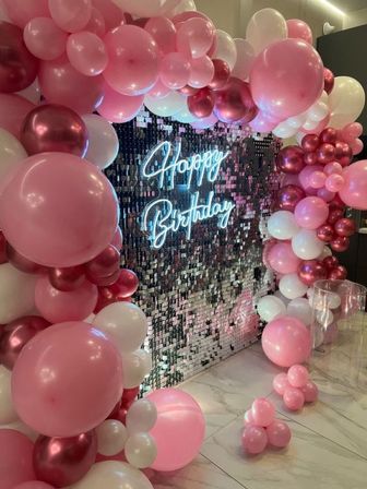 Festive indoor pink, white, and rose-gold balloon arch framing a sparkling sequin backdrop with a glowing neon 'Happy Birthday' sign — stylish birthday photo wall on marble floor.