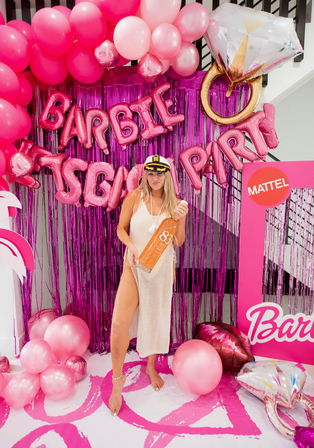 Woman in captain’s hat and white mesh cover-up posing with oversized bottle at a pink-themed indoor party photo booth with balloon arch, metallic fringe backdrop, foil letter balloons and giant diamond ring balloon