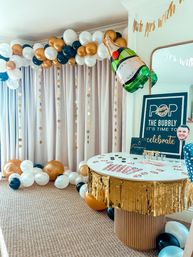 Indoor celebration setup with gold, white and black balloon arch and floor balloons, oversized champagne foil balloon, hanging gold confetti garlands and a round table with gold fringe skirt and party signage