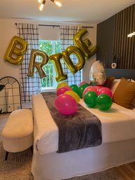 Cozy modern bedroom decorated for a bridal celebration with gold foil "BRIDE" letter balloons and colorful pink, green, and yellow balloons scattered on a white bed beneath plaid curtains