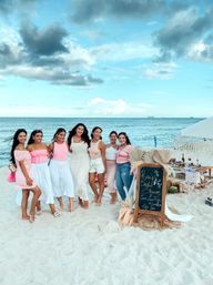 Seven women in pastel outfits posing on a sandy beach by a turquoise sea next to a boho bridal shower picnic setup and chalkboard sign.