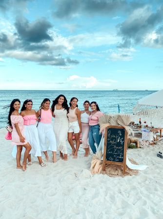 Seven women in pastel outfits posing on a sandy beach by a turquoise sea next to a boho bridal shower picnic setup and chalkboard sign.
