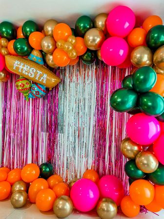 Colorful fiesta party photo backdrop with hot-pink, orange, green and gold balloons forming an arch around metallic pink-and-silver tinsel, accented by a foil ‘Fiesta’ sombrero and maraca balloon.