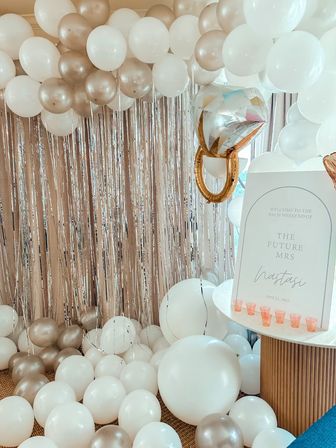Indoor bachelorette party decor with white and champagne-gold balloon arch and floor clusters, metallic fringe photo backdrop, oversized gold ring balloon, and a welcome sign on a marble-top table.
