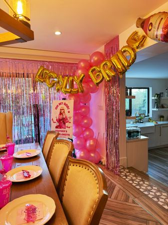 Indoor home dining room decorated for a pink-and-gold bachelorette: wooden table with place settings and pink cups, metallic fringe backdrop, pink balloon column and gold letter balloons reading 'LEGALLY BRIDE' near an open-plan kitchen.