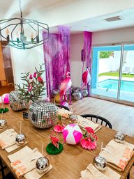 Vibrant pink flamingo poolside party setup in a dining room by sliding glass doors overlooking a backyard pool — wooden table with mirrored disco-ball centerpieces, pink-and-white beach ball, flamingo cutouts, metallic balloons and pink fringe backdrop.