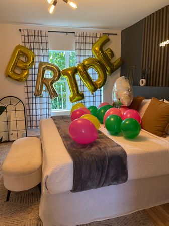 Cozy bedroom decorated for a bridal celebration: gold foil 'BRIDE' balloons above a bed with colorful pink, green, and yellow balloons scattered on a white quilt and gray throw.