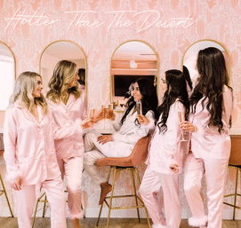 Five women in pink satin pajamas toasting champagne in a pink vanity-style bridal suite with arched mirrors — playful bachelorette pajama party scene.