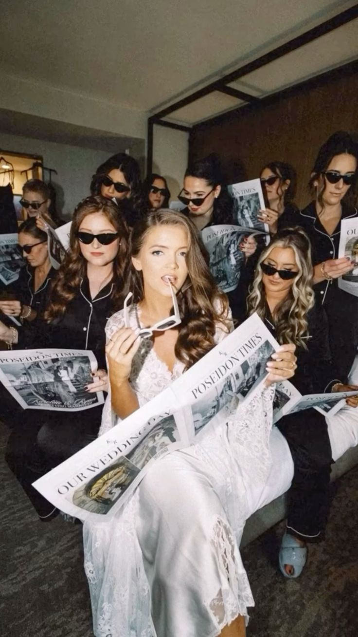 Bride in white lace robe lounging with bridesmaids in matching black pajamas and sunglasses, all reading wedding newspapers in a hotel suite