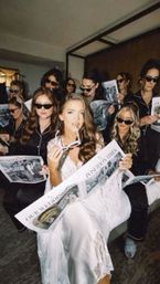 Bride in white lace robe lounging with bridesmaids in matching black pajamas and sunglasses, all reading wedding newspapers in a hotel suite