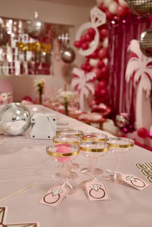 Pink bachelorette party table with gold-rimmed coupe glasses holding pink ping-pong balls, disco balls, balloon garland and palm cutout decorations in a festive indoor setting.