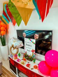 Colorful living room fiesta party décor for a bachelorette weekend: papel picado banners overhead, bright orange and pink balloons, a metallic ring balloon, TV with face cutouts, tabletop mini sombreros and cactus centerpieces around a welcome sign.
