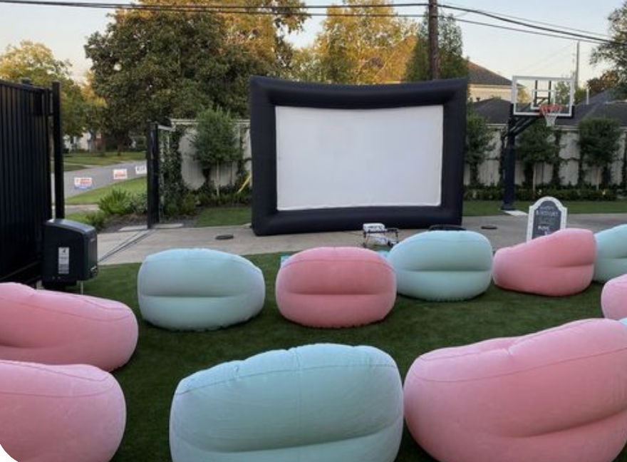 Cozy backyard outdoor movie setup with a large inflatable screen and pastel pink and mint inflatable beanbag seats arranged on a green lawn in a suburban yard