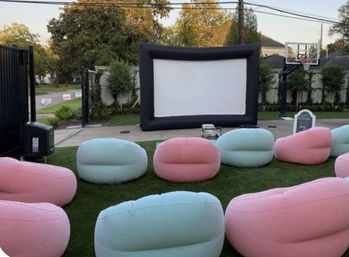 Cozy outdoor backyard movie setup with a large inflatable screen, projector on the grass, and pastel pink and mint inflatable beanbag seats arranged on a lawn near a basketball hoop in a suburban yard.
