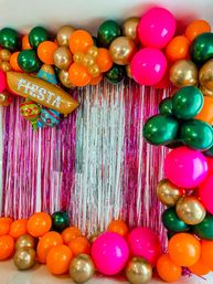 Fiesta-themed photo backdrop with pink metallic fringe, green, gold, orange and hot-pink balloon garland and inflatable FIESTA maracas