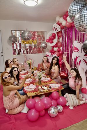 Group of women toasting at a pink-themed bachelorette party in a cozy apartment living room with balloon garlands, disco balls, fringe backdrop and charcuterie.
