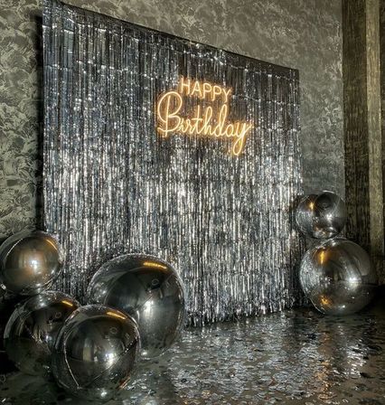 Glam indoor silver birthday photo backdrop with neon Happy Birthday sign, shimmering foil fringe curtain, oversized reflective silver balloons and confetti-strewn floor.