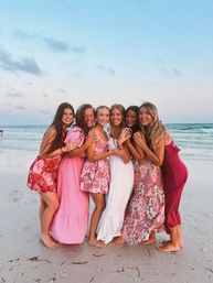 Six friends in colorful summer dresses hugging barefoot on a white-sand beach at sunset with gentle ocean waves and a pastel sky in the background.