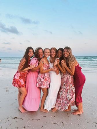 Six friends in colorful summer dresses hugging barefoot on a white-sand beach at sunset with gentle ocean waves and a pastel sky in the background.