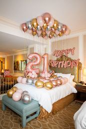 Festive rose gold, gold and silver balloons crowd a hotel bedroom ceiling and bed, featuring large glittery '21' number balloons and 'Happy Birthday' letter balloons over a decorated bed.