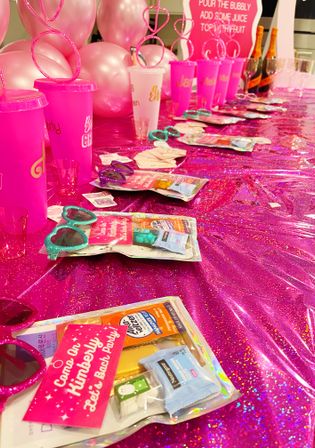 Indoor hot-pink party favor table for a bachelorette/girls' night with glitter tablecloth, rows of pink tumblers, heart-shaped sunglasses, packaged goodies, champagne bottles and pink balloons