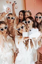 Bridal party selfie: women in white satin robes and white heart-shaped sunglasses pose with an instant camera against a pink floral backdrop, playful celebration.