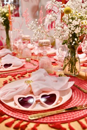 Romantic pink tablescape for Valentine’s Day with heart-shaped pink sunglasses on a plate, pale pink napkin with gold ring, gold flatware, woven pink placemat and baby’s breath flowers in a glass vase.