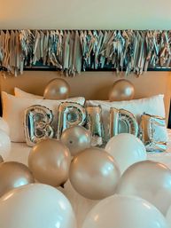 Silver 'BRIDE' foil balloons with pearl gold and white balloons scattered across a hotel bed beneath metallic tassel garland — bridal suite celebration decor
