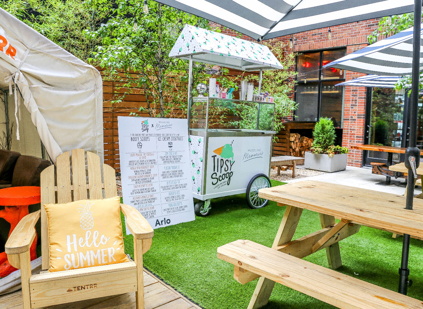 Sunny urban courtyard patio with wooden picnic tables and striped umbrellas, mobile ice cream cart and menu board on artificial turf, Adirondack chair with “Hello Summer” pillow and brick backdrop.