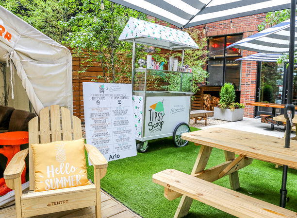Sunny urban courtyard patio with wooden picnic tables and striped umbrellas, mobile ice cream cart and menu board on artificial turf, Adirondack chair with “Hello Summer” pillow and brick backdrop.