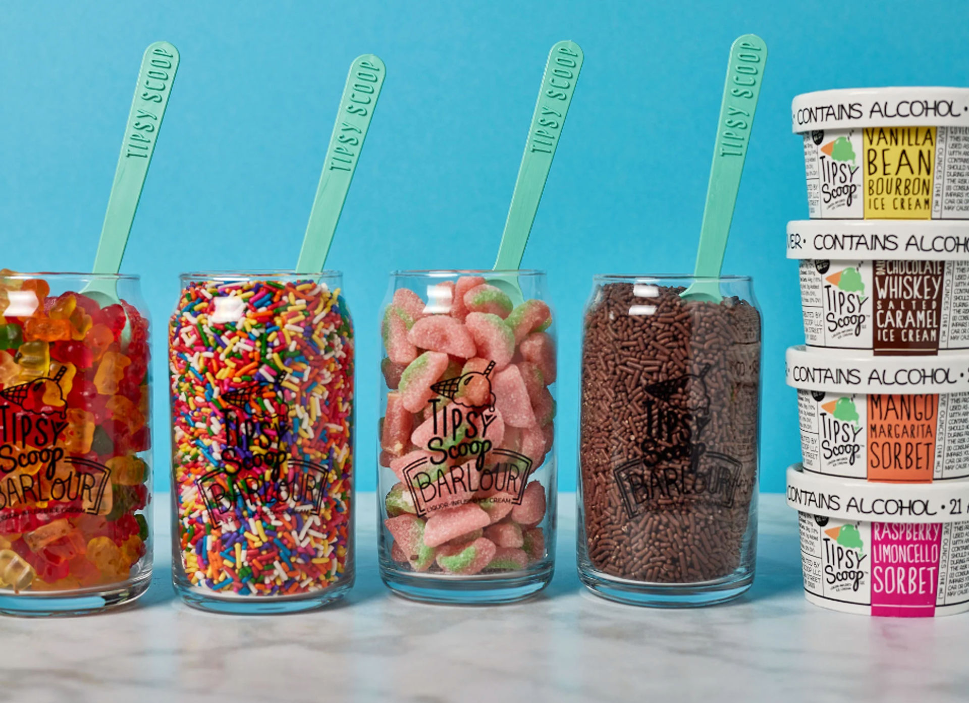 Four clear jars of ice cream toppings—gummy bears, rainbow sprinkles, sour peach candies, and chocolate jimmies—with mint-green spoons on a marble countertop and stacked pints of alcoholic ice cream against a bright blue background.