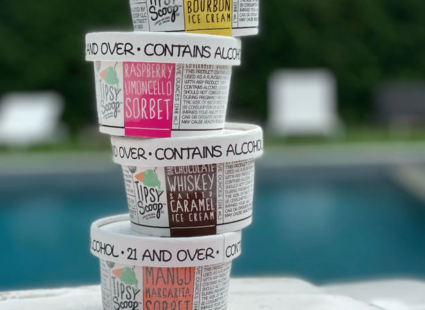 Stack of four small tubs labeled "Contains Alcohol" with flavors like raspberry limoncello sorbet, bourbon ice cream, whiskey salted caramel, and mango margarita sorbet stacked on a poolside ledge with a blurred backyard pool in the background.