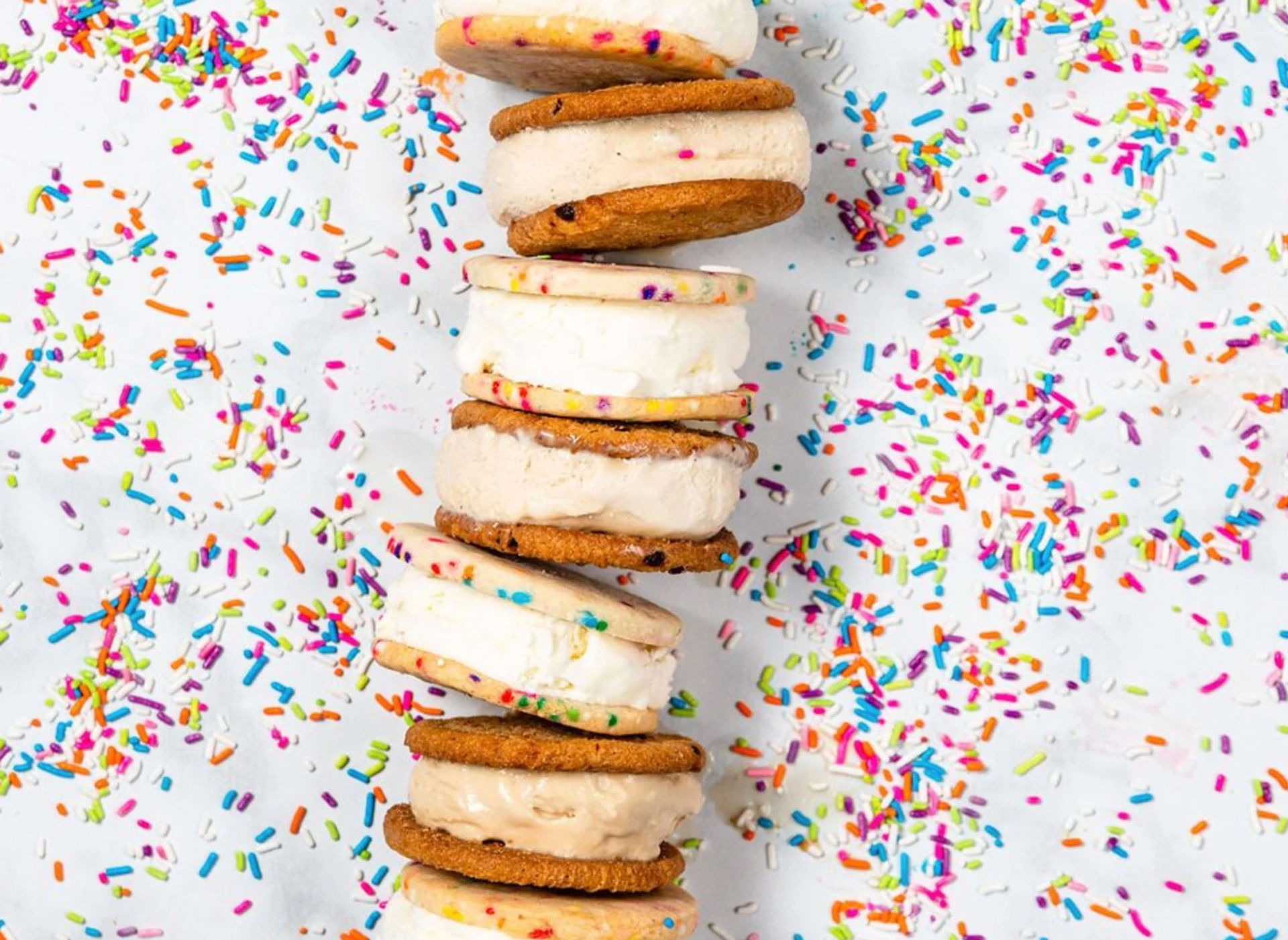 Stack of ice cream sandwich cookies with vanilla and caramel ice cream, surrounded by colorful rainbow sprinkles on a white background