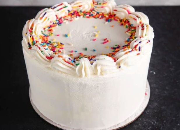 White buttercream layer cake with piped swirls and colorful rainbow sprinkles on top — simple birthday celebration cake on a dark countertop