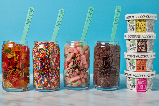 Four clear jars of ice cream toppings — gummy bears, rainbow sprinkles, peach sour candies, and chocolate sprinkles — each with a teal scoop, beside a stack of small pints labeled contains alcohol on a marble counter with a bright blue backdrop.