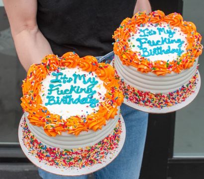 Two round birthday cakes with orange rosette frosting, blue handwritten 'It's My Fucking Birthday' message and rainbow sprinkles held by a person