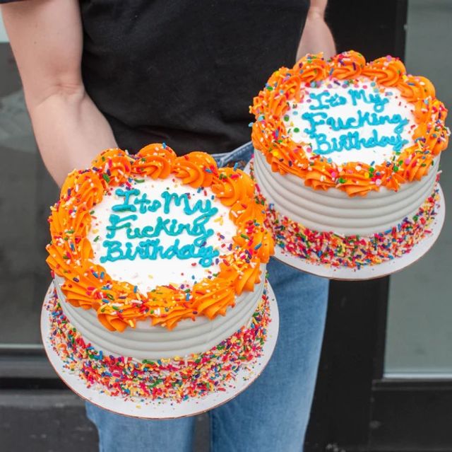 Two round birthday cakes with orange rosette frosting, blue handwritten 'It's My Fucking Birthday' message and rainbow sprinkles held by a person