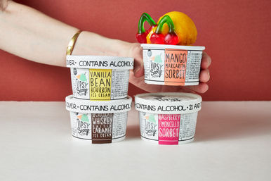 Bright studio shot of four boozy ice cream and sorbet cups with colorful flavor labels—vanilla bourbon, mango margarita, whiskey salted caramel and raspberry limoncello—stacked on a white tabletop against a red backdrop, one cup held with a mango scoop topped by gummy cherries.