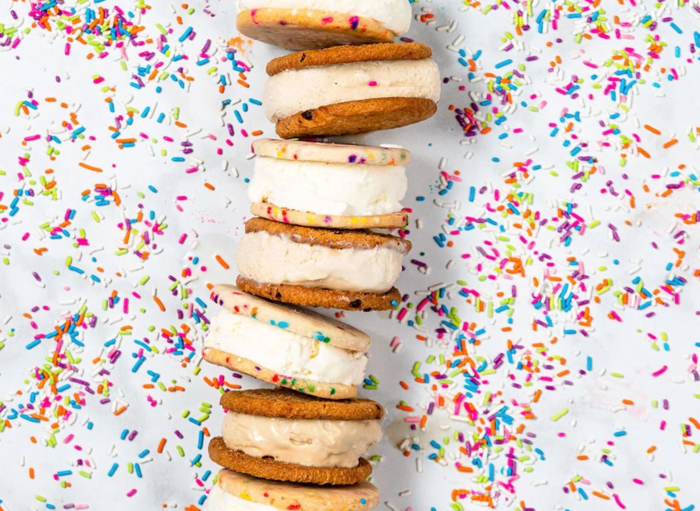 Playful stack of ice cream cookie sandwiches with vanilla and chocolate fillings between sprinkle-dotted cookies on a white background scattered with colorful rainbow sprinkles