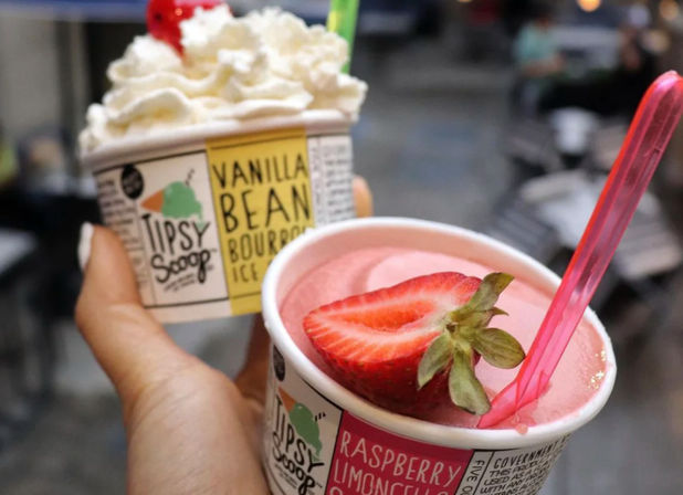 Hand holding two small cups of frozen treats: pink strawberry sorbet topped with a halved fresh strawberry and pink spoon, and vanilla-bean ice cream crowned with whipped cream and a cherry, blurred outdoor street background.
