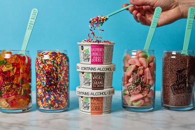 Stack of three alcohol‑labeled sorbet and ice cream pints with rainbow sprinkles being spooned on top, flanked by glass jars of multicolored sprinkles, gummy candies and chocolate jimmies with teal scoops on a marble surface and bright blue background.