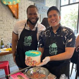 Two smiling staff in black aprons holding a blue-rosette celebration cake rolled in rainbow sprinkles at a bright neighborhood ice cream shop counter
