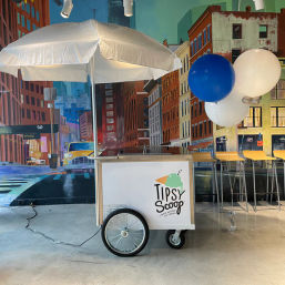 Playful indoor white ice cream cart with umbrella and blue-and-white balloons set against a colorful urban street mural, metal wheel and bar stools on a concrete floor.