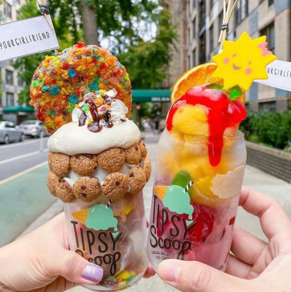 Two colorful over-the-top dessert drinks held on a city sidewalk — one cookie-studded milkshake with whipped cream, chocolate drizzle and a rainbow cereal cookie; the other a fruity sorbet float with mango scoops, red syrup, orange slice and a smiling sun garnish.