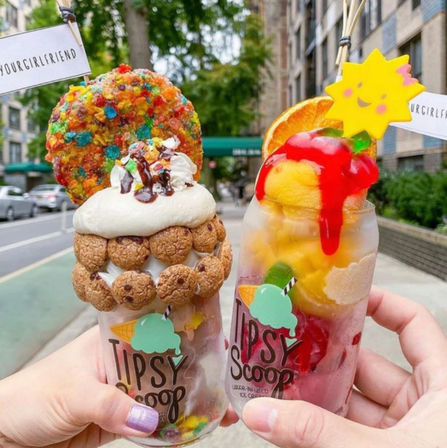 Two colorful over-the-top dessert drinks held on a city sidewalk — one cookie-studded milkshake with whipped cream, chocolate drizzle and a rainbow cereal cookie; the other a fruity sorbet float with mango scoops, red syrup, orange slice and a smiling sun garnish.