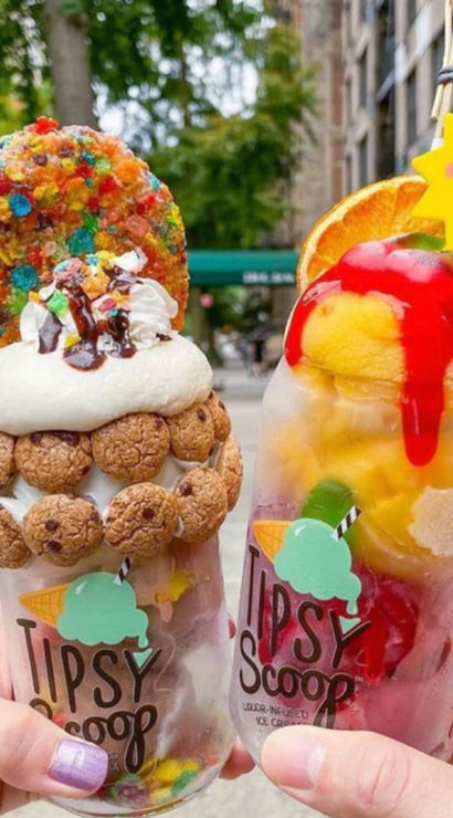 Two colorful over-the-top dessert drinks held on a city sidewalk — one cookie-studded milkshake with whipped cream, chocolate drizzle and a rainbow cereal cookie; the other a fruity sorbet float with mango scoops, red syrup, orange slice and a smiling sun garnish.