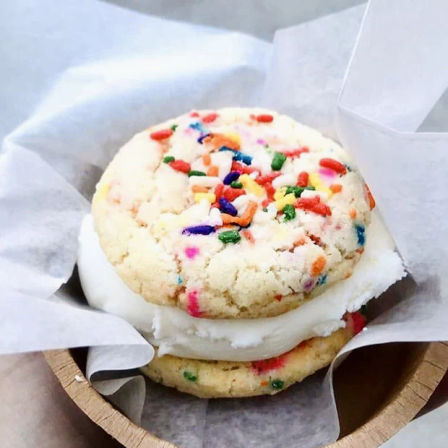 Close-up of a colorful funfetti sprinkle cookie ice cream sandwich with vanilla ice cream between two soft cookies, served on parchment paper in a small wooden bowl.