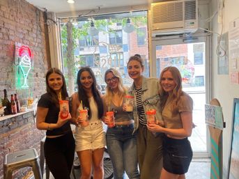 Five smiling friends inside an urban ice cream parlor holding tall, candy-topped milkshakes; exposed brick wall with neon ice cream sign and a large front window showing a tree-lined city street.