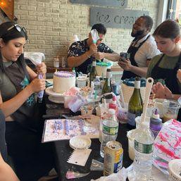 Four adults at a cake-decorating workshop in a bright café-style studio, piping pastel frosting onto layered cakes amid sprinkles, piping bags, bottles and decorating tools on a crowded table.