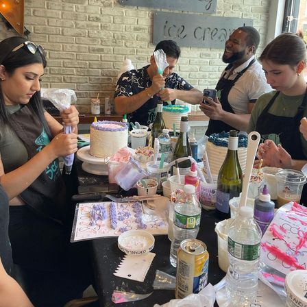 Four adults at a cake-decorating workshop in a bright café-style studio, piping pastel frosting onto layered cakes amid sprinkles, piping bags, bottles and decorating tools on a crowded table.